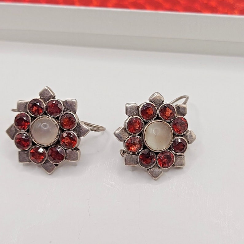 May include: A pair of silver earrings with a starburst design. Each earring features a central white pearl surrounded by 12 red gemstones.