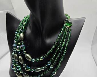 May include: A multi-strand green beaded necklace displayed on a black mannequin. The necklace features various bead shapes, including round and oval, with iridescent and metallic finishes. The necklace is a vintage style.