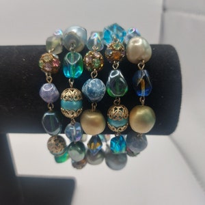May include: A multi-strand bracelet featuring blue, green, gold, and white beads. The beads are of various shapes and sizes, including round, oval, and square.