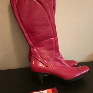 May include: A pair of knee-high, red leather boots with a pointed toe and a small, black heel. The boots have a glossy finish and a decorative seam detail. A price tag is attached.