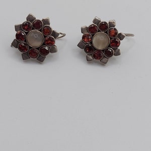 May include: A pair of silver earrings with a starburst design. Each earring has a central white stone surrounded by red gemstones.