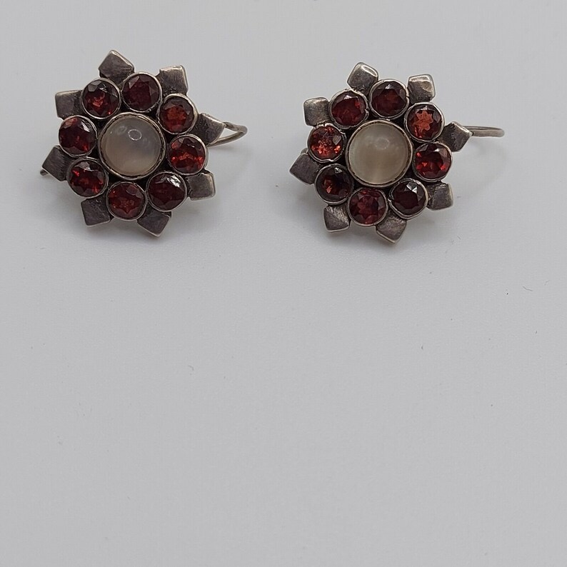 May include: A pair of silver earrings with a starburst design. Each earring has a central white stone surrounded by red gemstones.