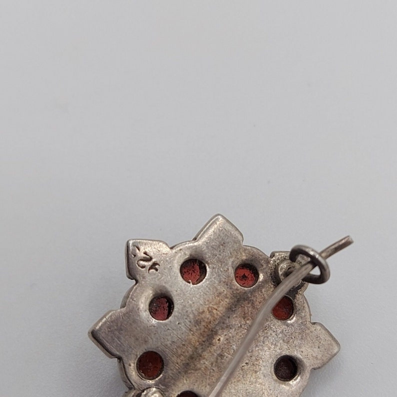 May include: Silver brooch with a starburst design and red stones. The back of the brooch has a silver pin and a clasp. The brooch is marked "925" on the back.