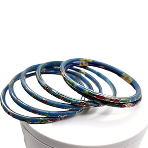 May include: A collection of five blue enamel bangles with floral designs in red, green, and yellow. The bangles are stacked on a white surface, showcasing their intricate patterns and vibrant colors. The jewelry is a classic accessory.