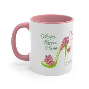Puede incluir: Taza de café de cerámica blanca con borde y asa de color rosa. La taza presenta un zapato de tacón alto verde con rosas rojas y el texto "Alpha Kappa Alpha 1908".