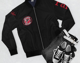 May include: Black bomber jacket with a red Atlanta Falcons logo on the front and "Rise Up" and "Falcons" in red on the sleeves. The jacket is zipped up and laid out on a white surface with a pair of black jeans and white trainers.