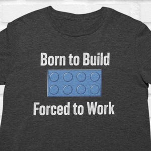 May include: Dark grey t-shirt with the text "Born to Build Forced to Work" in white. A blue toy brick graphic is centered between the text. The shirt is made of a soft material.