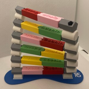 Könnte beinhalten: Ein 3D-gedrucktes Modell einer DNA-Doppelhelix. Das Modell besteht aus bunten Plastikblöcken mit den Namen der vier Stickstoffbasen: Adenin, Cytosin, Guanin und Thymin. Das Modell ist auf einem blauen Sockel montiert.