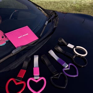 May include: A collection of car accessories, including heart-shaped and oval-shaped pull straps in various colors: red, pink, black, and purple. The image also shows a pink package with the text "SHIFTY STICKERS". The items are arranged on a dark surface.