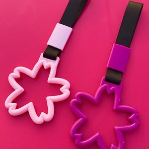 May include: Two flower-shaped car charms in pink and purple, each with a black strap and a matching colored clasp. The charms are set against a vibrant pink background.