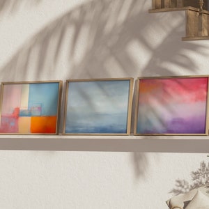 May include: Three abstract paintings in wooden frames hang on a white wall above a shelf. The paintings are in shades of blue, orange, and pink. The paintings are in a living room setting with a wicker basket and a potted plant.