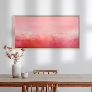 Può includere: Un dipinto astratto incorniciato, con una palette di colori rosa e rossi. L'opera d'arte è esposta sopra un tavolo di legno, con un vaso bianco di fiori secchi e una piccola tazza bianca. La cornice è in legno chiaro.