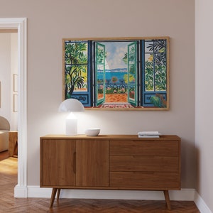Puede incluir: Una c&oacute;moda de madera con dos puertas y tres cajones. La c&oacute;moda es de color marr&oacute;n claro y tiene un dise&ntilde;o moderno. Una l&aacute;mpara blanca y un cuenco est&aacute;n encima de la c&oacute;moda. Un cuadro enmarcado de una ventana con vista a un balc&oacute;n y el mar est&aacute; colgado en la pared detr&aacute;s de la c&oacute;moda.