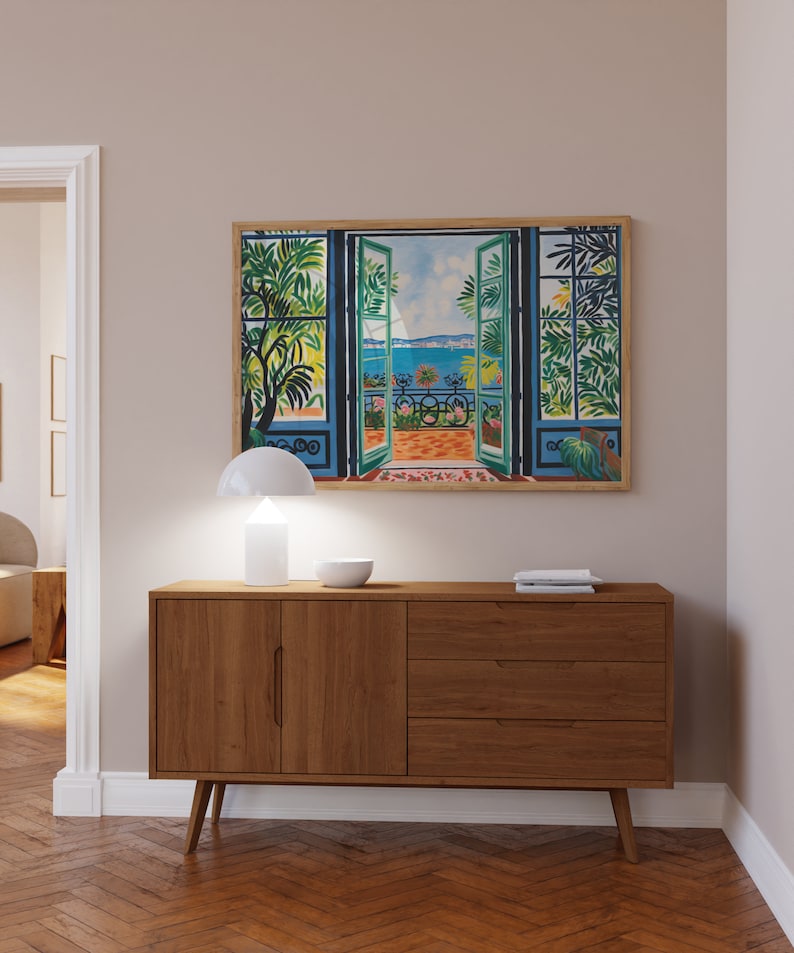 Puede incluir: Una c&oacute;moda de madera con dos puertas y tres cajones. La c&oacute;moda es de color marr&oacute;n claro y tiene un dise&ntilde;o moderno. Una l&aacute;mpara blanca y un cuenco est&aacute;n encima de la c&oacute;moda. Un cuadro enmarcado de una ventana con vista a un balc&oacute;n y el mar est&aacute; colgado en la pared detr&aacute;s de la c&oacute;moda.