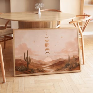 Può includere: Una stampa artistica da parete rettangolare incorniciata raffigurante un paesaggio desertico con cactus saguaro, colline e un sentiero tortuoso sotto un cielo che mostra una sequenza verticale di fasi lunari, stelle e un sole al tramonto.