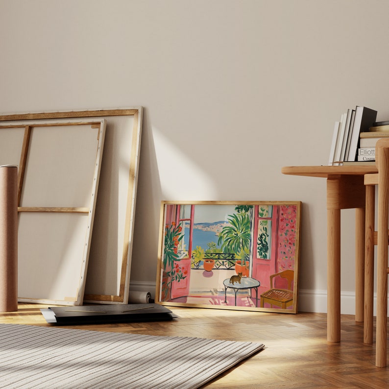 Puede incluir: Una impresi&oacute;n enmarcada de una pintura que representa una vista desde una ventana con un balc&oacute;n. El balc&oacute;n tiene una mesa peque&ntilde;a y sillas, y un gato est&aacute; sentado en la mesa. La vista desde la ventana es de un cuerpo de agua azul y colinas verdes en la distancia. La pintura est&aacute; en un marco de madera claro.