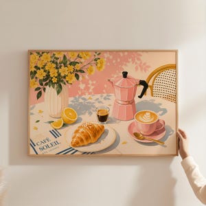 Könnte beinhalten: Ein gerahmter Wandkunstdruck zeigt eine Frühstücksszene mit einer rosa Mokkakanne, einer Tasse Latte mit Milchschaum, einem Croissant auf einem Teller, einem Glas Espresso, einer Vase mit gelben Blumen und einer Zitrone, mit dem Text CAFÉ SOLEIL BON JOURNÉE.