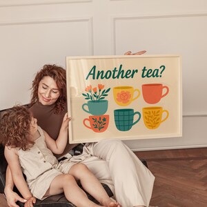 May include: A framed print featuring the words "Another tea?" and six illustrated teacups in various colours. The teacups are teal, yellow, orange, and green, each with a unique floral or patterned design. The print is held by a person and a child.
