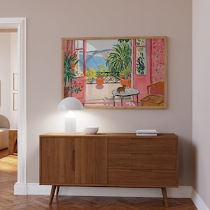 Puede incluir: Un aparador de madera con dos puertas y tres cajones, una l&aacute;mpara blanca y un cuadro enmarcado de un balc&oacute;n con vistas a una masa de agua azul. La pintura representa un gato sentado en una mesa con una planta en maceta y una silla.