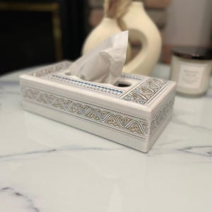 May include: A white rectangular tissue box with gold and blue geometric patterns. The box is on a marble-effect surface, with tissues visible. A candle and a decorative vase are in the background.