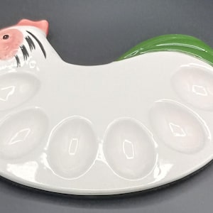 May include: A white ceramic egg serving tray shaped like a chicken. The tray features a pink head with a yellow beak, black eye details, and a green tail section. It includes six oval-shaped indentations designed to hold individual eggs securely.