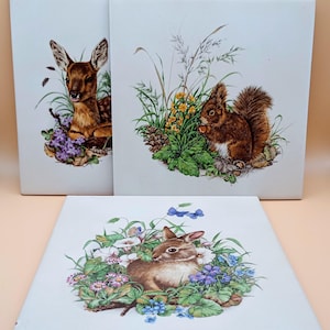May include: Three ceramic tiles with a white background and a brown border. Each tile features a different animal surrounded by flowers and greenery. The first tile features a fawn, the second tile features a squirrel, and the third tile features a rabbit.