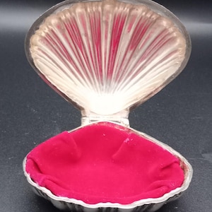 May include: A silver-toned metal clam shell-shaped trinket box with a hinged lid. The interior of the bottom half is lined with a vibrant, deep red velvet fabric. The interior of the lid features a ribbed, metallic finish mimicking a natural shell.