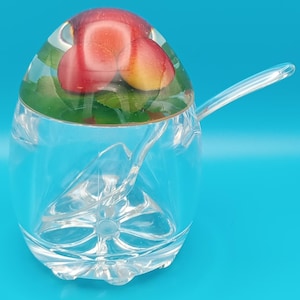 May include: A clear glass sugar bowl with a lid containing red and yellow fruit and green leaves. A clear spoon rests inside the bowl. The bowl sits on a blue surface.