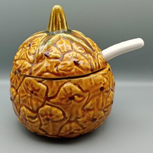 May include: A ceramic pineapple-shaped serving dish with a lid and a white spoon. The dish is a warm brown colour with darker brown accents and a textured surface. The lid has a green and brown stem.
