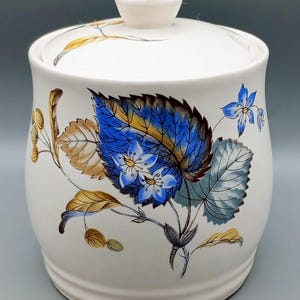 May include: White ceramic sugar bowl with a lid. The bowl features a floral design with blue, yellow and brown leaves and flowers.