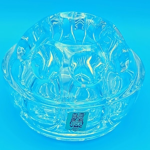 May include: A clear glass flower frog, designed for arranging flowers, with a rounded shape and multiple holes. The frog has a textured surface and a sticker that reads "VMC FRANCE". It is set against a blue background.