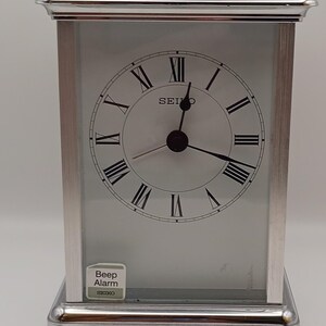 May include: A silver Seiko alarm clock with a rectangular design. The clock face is white with black Roman numerals and hands. It features a handle and a label that reads "Beep Alarm".