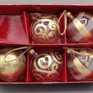 May include: A set of five spherical Christmas baubles in a red cardboard box. The collection includes one solid gold glitter bauble, two clear glass baubles with gold swirl patterns, and two clear glass baubles with gold and red plaid stripes.