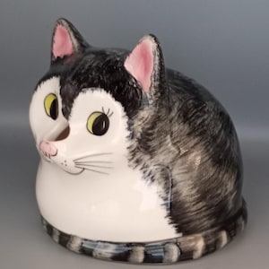May include: A ceramic cat-shaped figurine with a glossy finish. The cat has a white chest and face, with black and grey tabby markings on its back and head. It features pink-tinted ears, yellow eyes with black pupils, and a small pink nose.