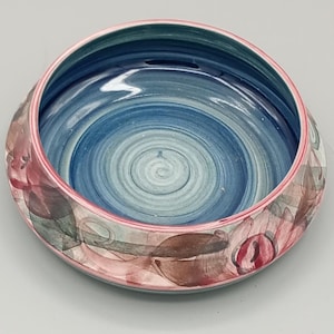 May include: A round ceramic bowl featuring a smooth, glossy interior with concentric blue and teal circular brushstroke patterns. The exterior rim is painted with a thin pink line, while the outer body displays a soft, abstract floral motif.