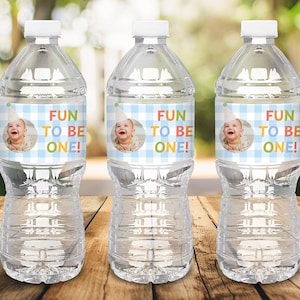 Puede incluir: Tres botellas de agua de plástico transparente con tapas blancas. Cada botella tiene una etiqueta con el texto "FUN TO BE ONE!" y una foto de un bebé con un gorro de fiesta. Las etiquetas tienen un patrón de cuadros azules y blancos.