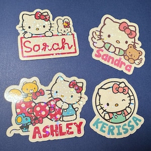 May include: Four Hello Kitty themed stickers with glitter accents. Each sticker features a different Hello Kitty character and a name: Sarah, Sandra, Ashley, and Kerissa.