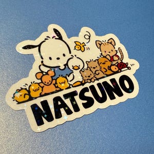 Puede incluir: Una pegatina blanca con un perro de dibujos animados y otros animales. La pegatina tiene un acabado brillante y dice "NATSU NO" en letras negras.