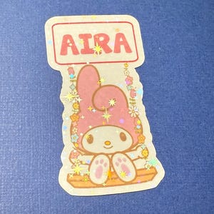 May include: A pink Hello Kitty character with a red sign that says "Aira" above her. She is sitting on a swing with flowers and stars around her. The sticker is glittery.