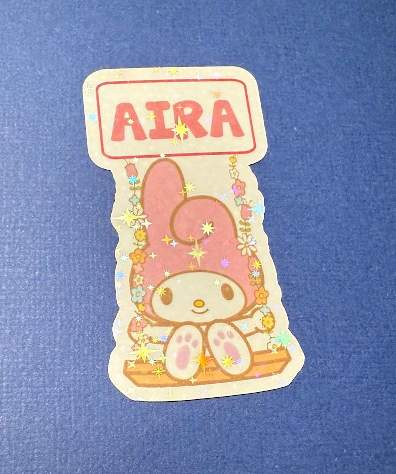 May include: A pink Hello Kitty character with a red sign that says "Aira" above her. She is sitting on a swing with flowers and stars around her. The sticker is glittery.