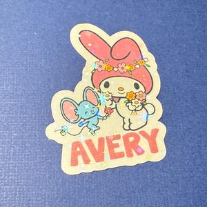 May include: A pink and white Hello Kitty sticker with a blue mouse character. The sticker has a glittery finish and says "AVERY" in red letters.