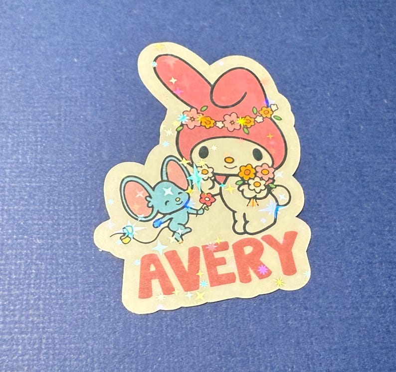 May include: A pink and white Hello Kitty sticker with a blue mouse character. The sticker has a glittery finish and says "AVERY" in red letters.