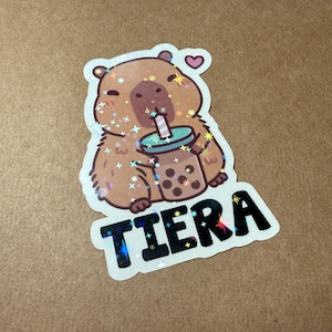 May include: A sticker featuring a cute capybara drinking boba tea. The capybara is brown with a pink nose and a small pink heart above its head. The word "TIERA" is written in black letters with a holographic effect.