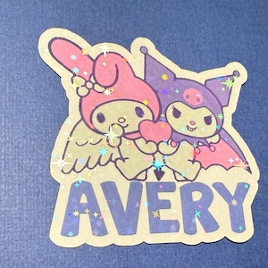 May include: A sticker featuring My Melody and Kuromi, two popular Sanrio characters. My Melody is a pink rabbit with angel wings, and Kuromi is a black cat with bat wings. They are holding a heart together. The sticker has a glittery finish and says "AVERY" in blue letters.