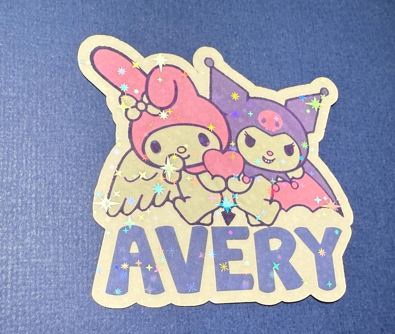 May include: A sticker featuring My Melody and Kuromi, two popular Sanrio characters. My Melody is a pink rabbit with angel wings, and Kuromi is a black cat with bat wings. They are holding a heart together. The sticker has a glittery finish and says "AVERY" in blue letters.