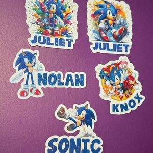 May include: Five colorful stickers featuring the Sonic the Hedgehog character. The stickers have different designs and include the names "JULIET", "NOLAN", "KNOX", and "SONIC".