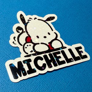 Puede incluir: Una pegatina blanca con un perro de dibujos animados en blanco y negro y el nombre "Michelle" en letras negras sobre un fondo brillante.