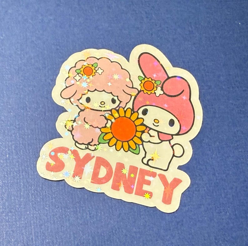 May include: A pink and white sticker with a cartoon bunny and a sheep holding a sunflower. The sticker says "SYDNEY" in pink letters with white stars.