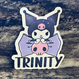 May include: A holographic sticker featuring a cartoon character with a black head, purple bow, and a pink skull. The word "TRINITY" is written in silver letters at the bottom. The sticker has a glittery, iridescent finish.
