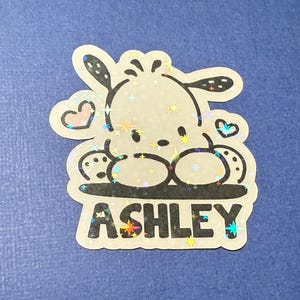 Puede incluir: Una pegatina hologr&aacute;fica con un perro de dibujos animados sobre un fondo brillante. El perro est&aacute; sentado con las patas cruzadas y tiene un coraz&oacute;n sobre su cabeza. La pegatina dice "ASHLEY" en letras negras.
